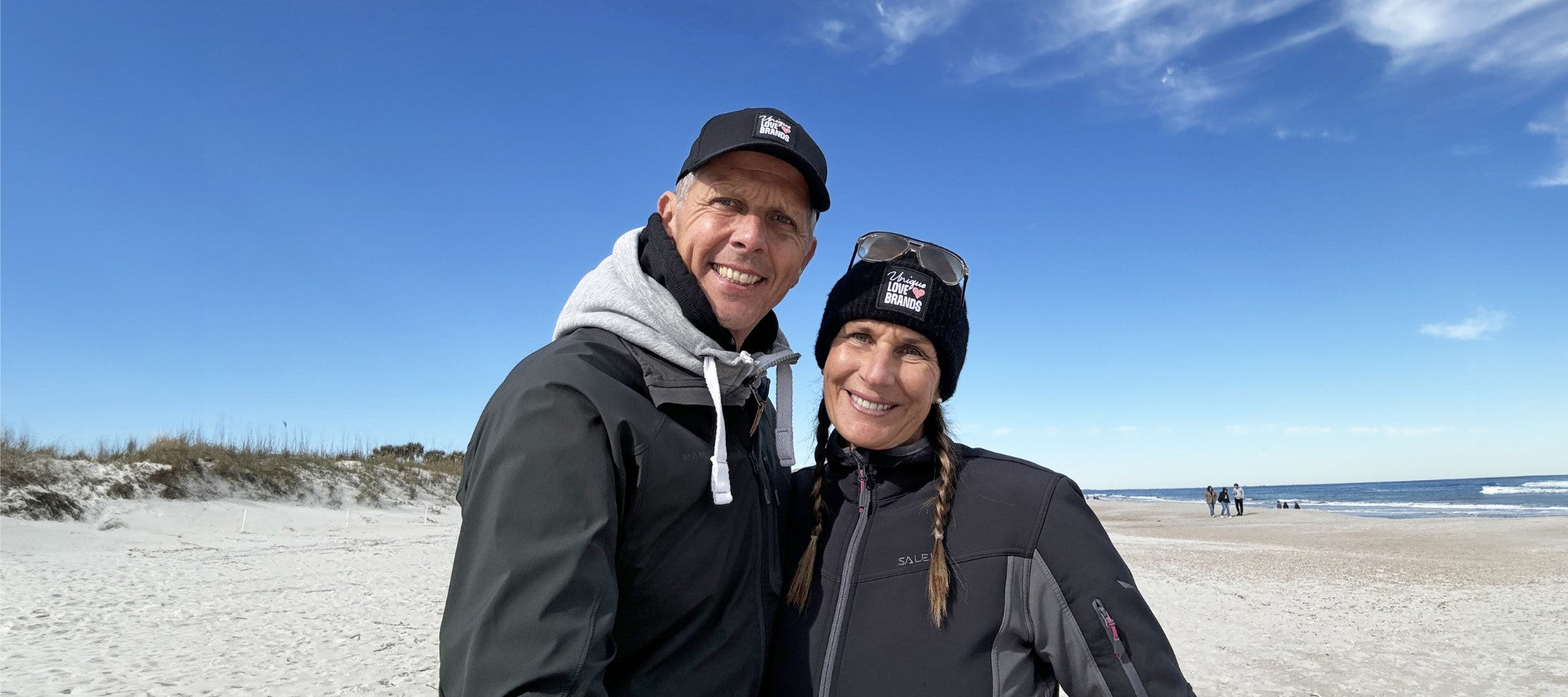 A woman and a man at the beach wearing a black cap and a beanie from unique love brands