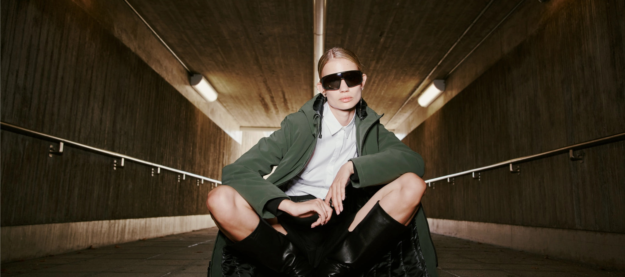 A blond woman wearing a white blouse, black boots, and a green jacket cowering on the ground